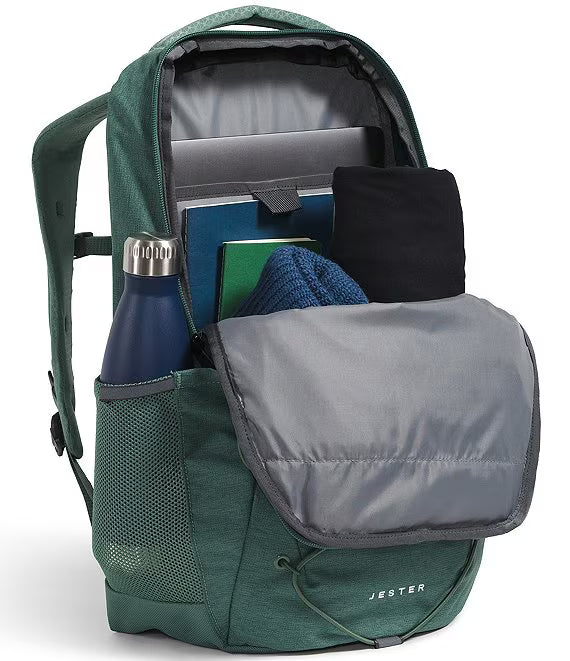 A green Jester backpack shown open, filled with travel items including a blue water bottle, notebook, folded black garment, and blue knit item, on a clean white background.