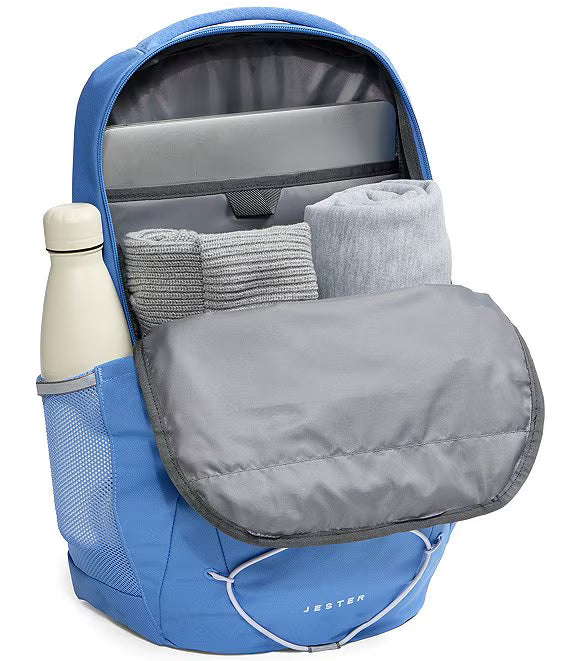 A blue Jester backpack shown open, revealing a gray padded compartment, a laptop sleeve, rolled towels, and a side mesh bottle holder with a white bottle, on a white background.