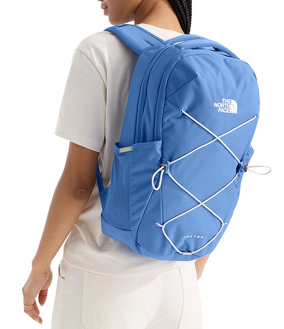 A person wearing a white t-shirt and white pants, shown from the back and side, carrying a blue The North Face backpack with white bungee cords on a white background.