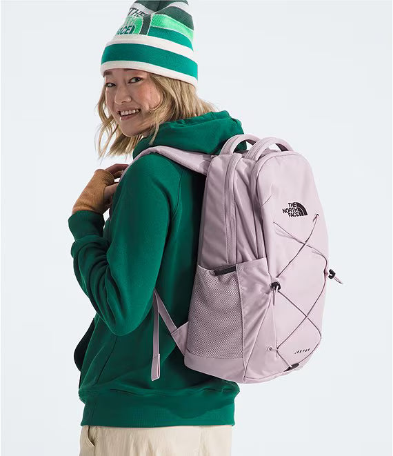 A person wearing a green hoodie and a green-and-white beanie carries a light pink The North Face backpack against a plain light gray studio background.