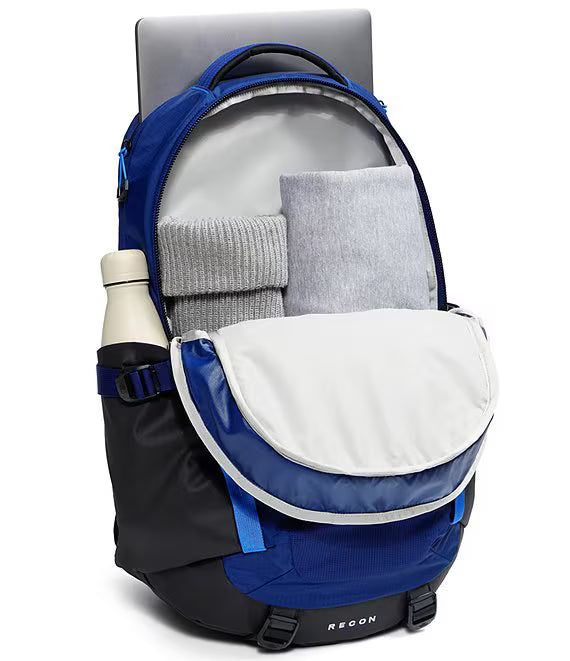 A blue and black REEGN backpack shown open, with a gray laptop, folded gray sweater, folded gray blanket, and a white bottle in the side pocket, on a white background.