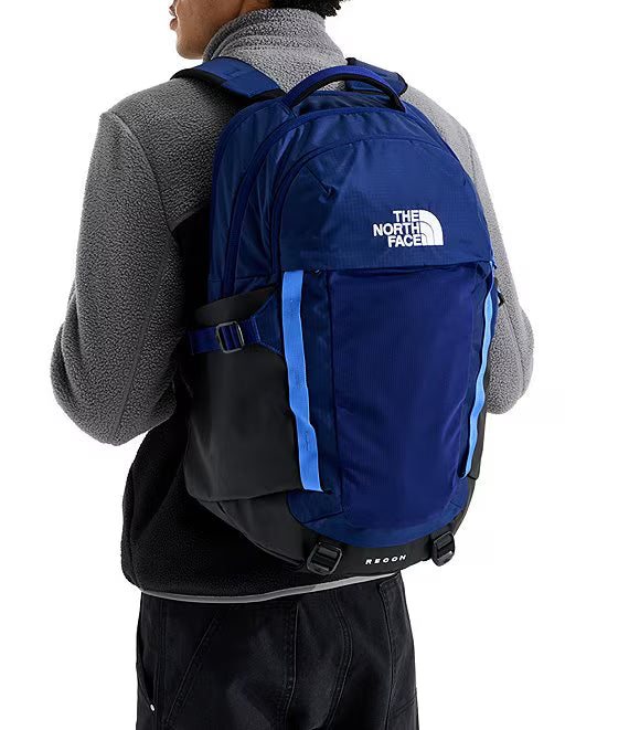 A person wearing a gray fleece jacket and black pants, shown from the back while wearing a blue and black The North Face backpack with light blue accents.