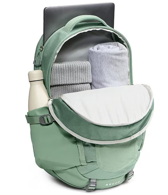 A sage green backpack shown open, containing a gray laptop, rolled gray sweater, light gray blanket, and a cream water bottle in its side pocket. The brand name 'RECOLL' is visible on the lower front.