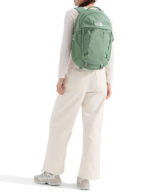 A person shown from the back wearing a sage green The North Face backpack, cream pants, and light-colored sneakers against a plain white studio background.
