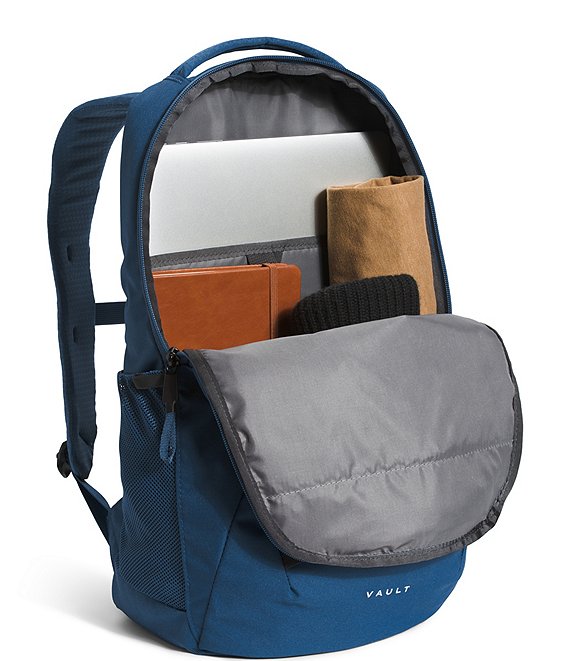 A navy blue backpack shown open, revealing its interior storage compartments with a laptop, notebook, rolled item, and hat. The front pocket is open, showing a gray padded interior, and the brand name 'VAULT' is printed in white near the bottom right corner.