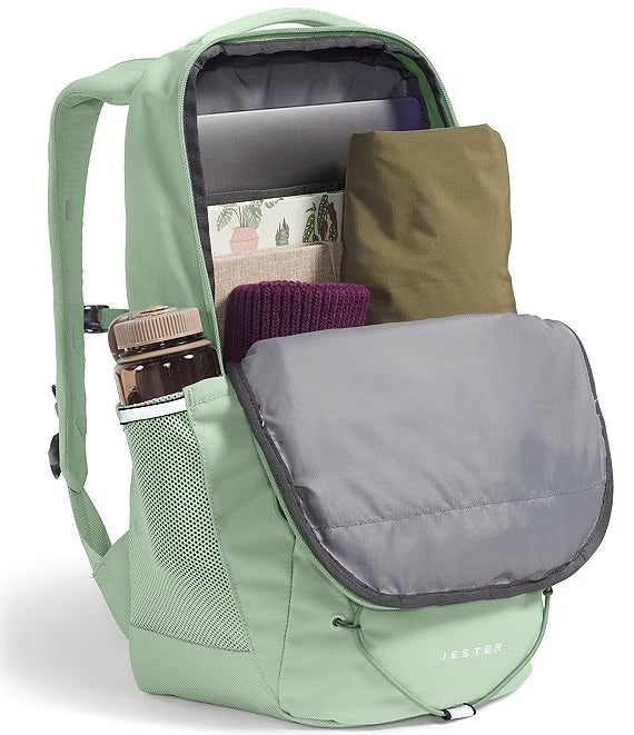 A sage green Jester backpack shown open, displaying its interior storage compartments filled with a laptop, pouches, and accessories. The backpack features a side mesh pocket with a water bottle and has the Jester brand name subtly printed on the lower front.