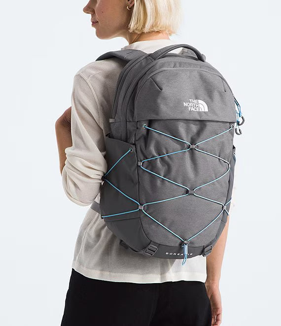 A woman wearing a light gray long-sleeve shirt and black pants is shown from the back, carrying a dark gray The North Face Borealis backpack with light blue bungee cords on a white studio background.