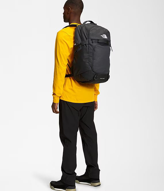 A man wearing a bright yellow long-sleeve shirt, black pants, and black sneakers is shown from the back, wearing a dark gray backpack by The North Face. The photo is taken against a clean, light gray studio background.