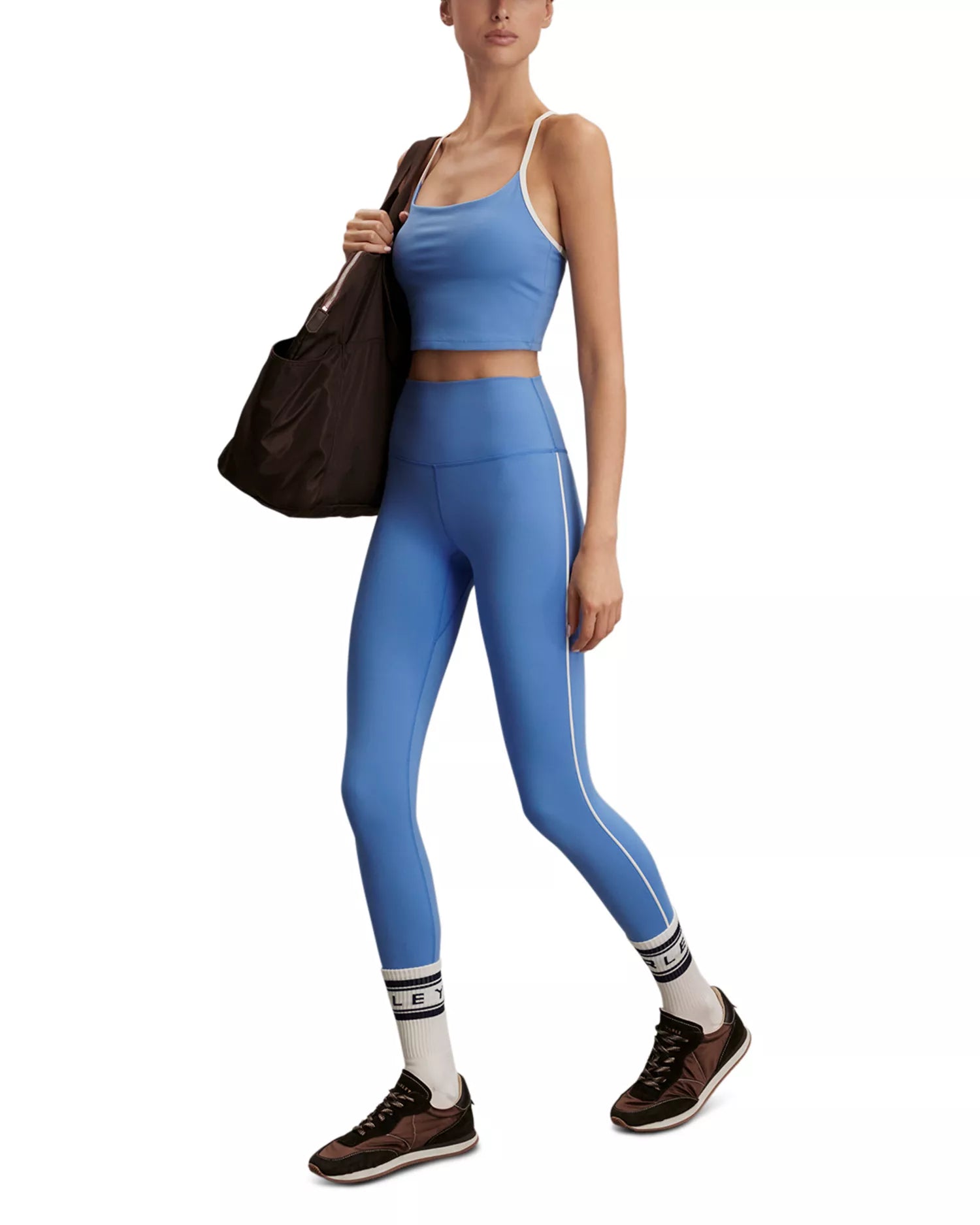 A woman modeling a matching athletic set consisting of a light blue sports bra and high-waisted leggings with white side stripes, styled with brown sneakers, white socks, and a dark brown tote bag against a plain white background.