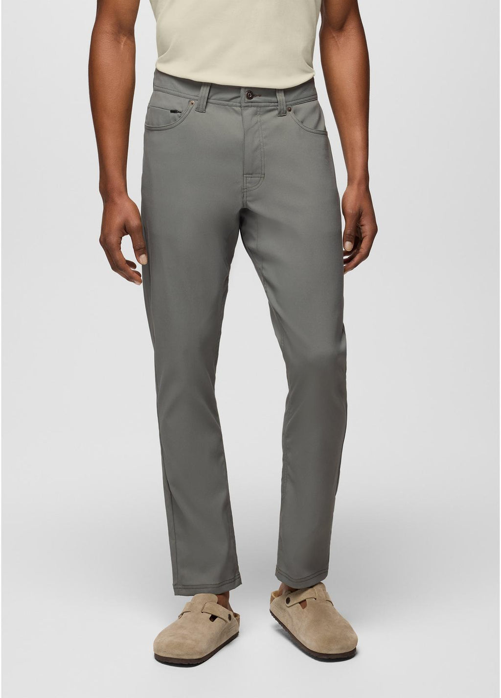 A person wearing slim-fit gray trousers, paired with a light beige top and tan clogs, shown from the waist down against a light gray studio background.