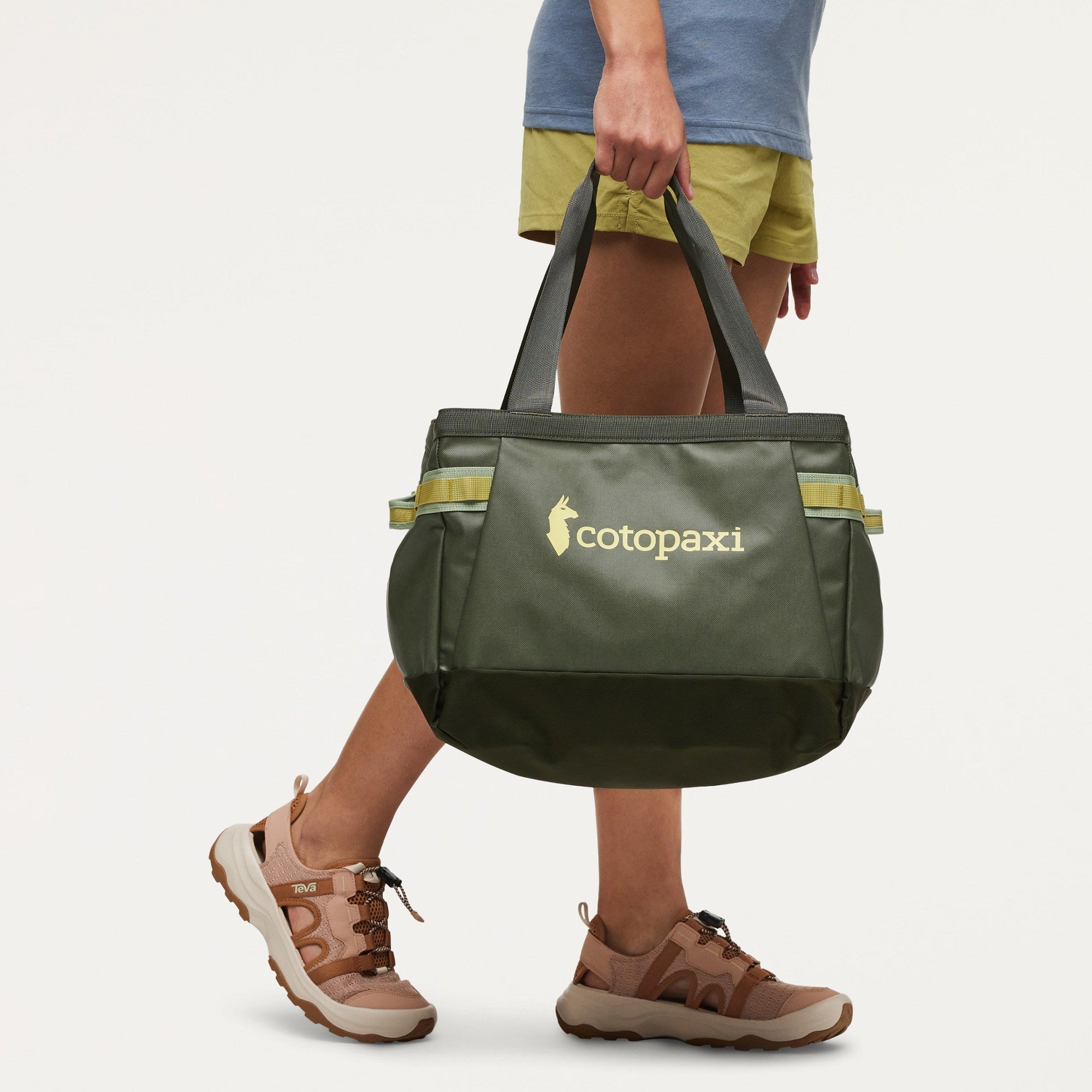 A person carrying a large Cotopaxi insulated tote bag in olive green with yellow-green reflective accents and a gold logo, shown against a clean off-white studio background.