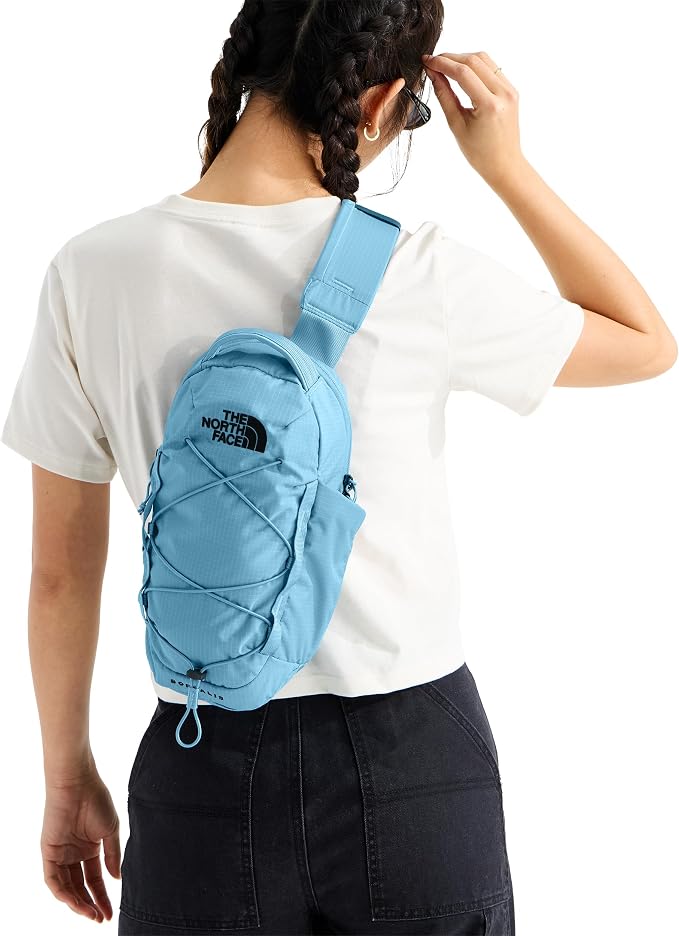 A woman from behind wearing a light blue sling backpack by The North Face over a white t-shirt and dark jeans, against a plain white background.