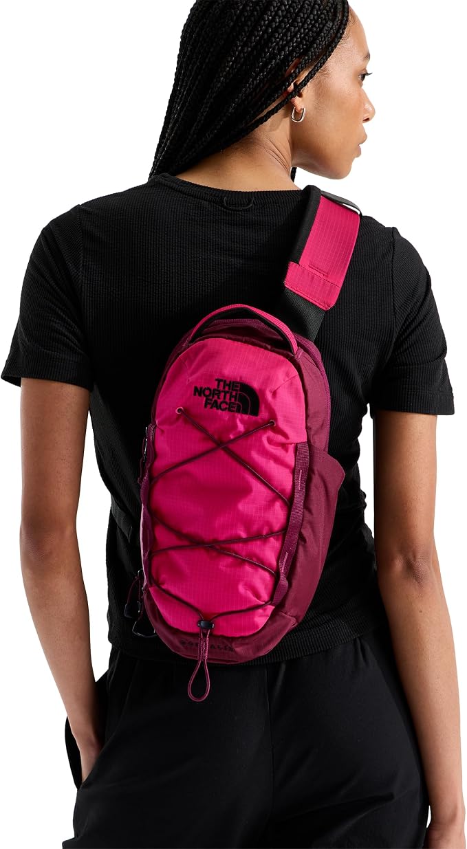 A woman wearing a black t-shirt and black pants, viewed from the back, carrying a bright pink sling backpack by The North Face. The backpack has a wide shoulder strap and a front zippered pocket with crisscross compression straps.