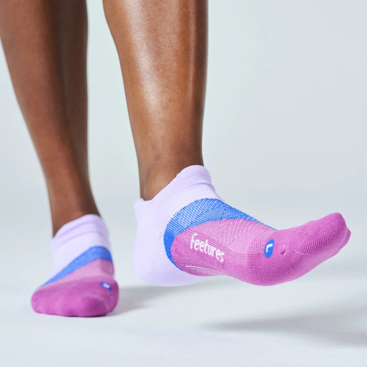 A close-up of a person's lower legs wearing low-cut athletic socks. The socks are primarily light purple with a bright pink toe and sole, a blue mesh side panel, and the brand name 'feetures' printed in white on the side.