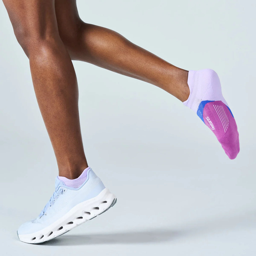 A person wearing light purple no-show socks with a blue and pink sole, paired with white running shoes, captured mid-step against a light gray studio background.