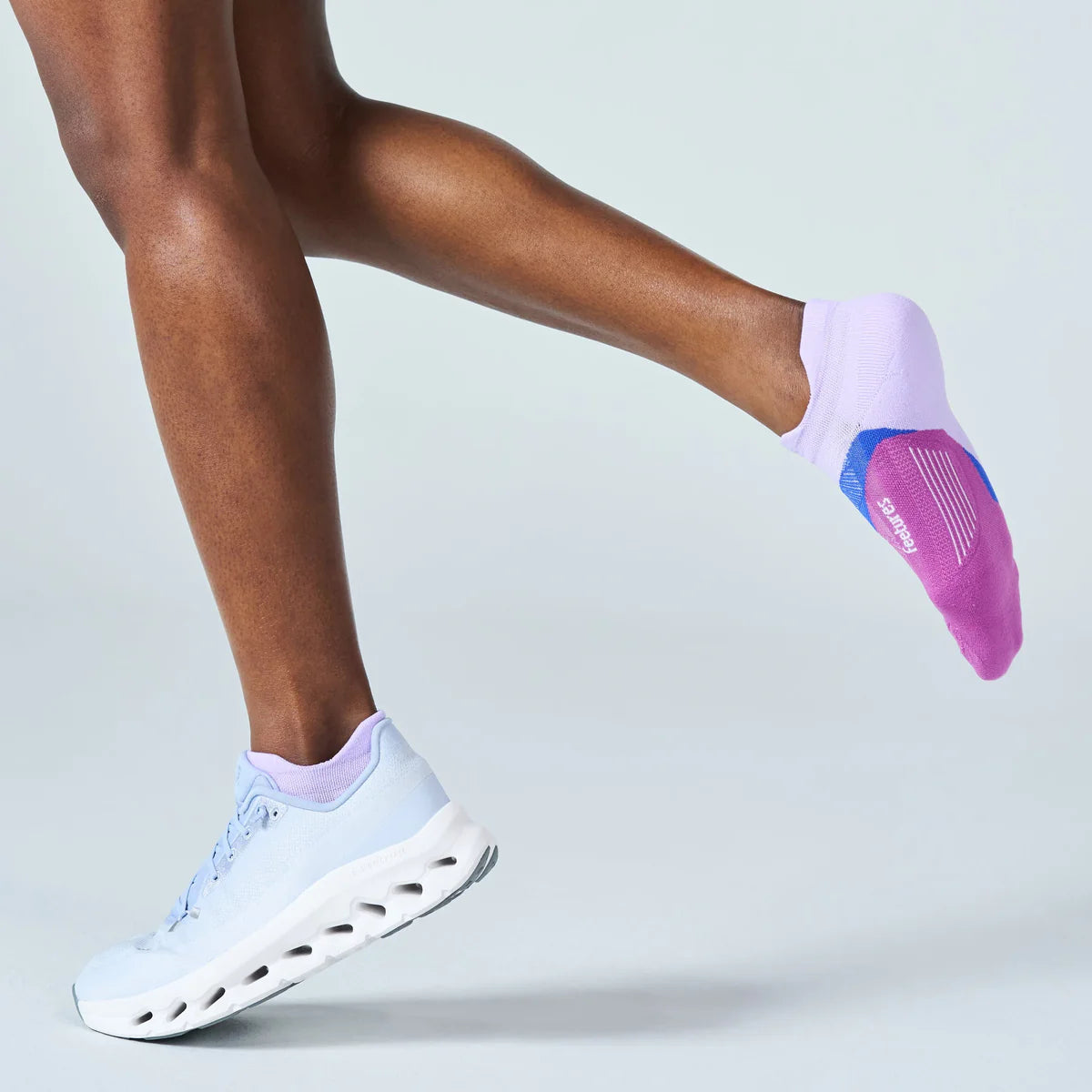 A person wearing light purple no-show socks with a blue and pink sole, paired with white running shoes, captured mid-step against a light gray studio background.