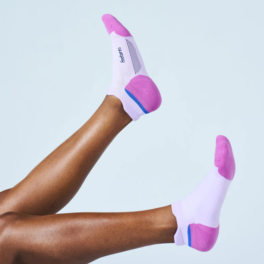 A person's legs raised in the air wearing white low-cut athletic socks with pink toes, heels, and heels, and a blue stripe near the cuff. The brand name 'feetrs' is printed on the top of one sock.