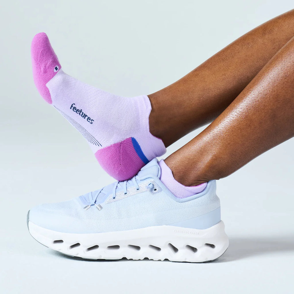 A person wearing light blue athletic sneakers and white socks with pink and purple accents. The sock has the brand name 'feetures' printed on the side, shown against a clean light gray studio background.