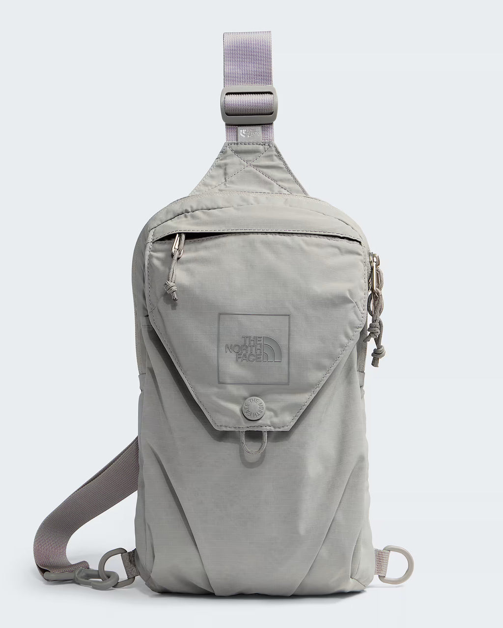 A light gray sling backpack by The North Face, featuring a front flap pocket with a snap button closure, a top zipper, and an adjustable shoulder strap, shown against a light gray background.