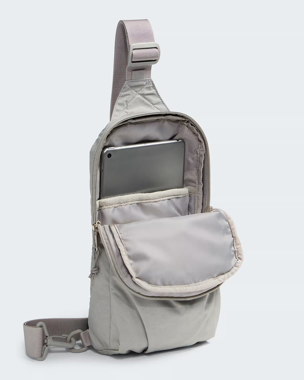 A light gray nylon sling backpack shown open, with a tablet inserted into the top compartment and a front flap pocket. The bag has a wide adjustable shoulder strap and a detachable crossbody strap, photographed on a clean light gray background.