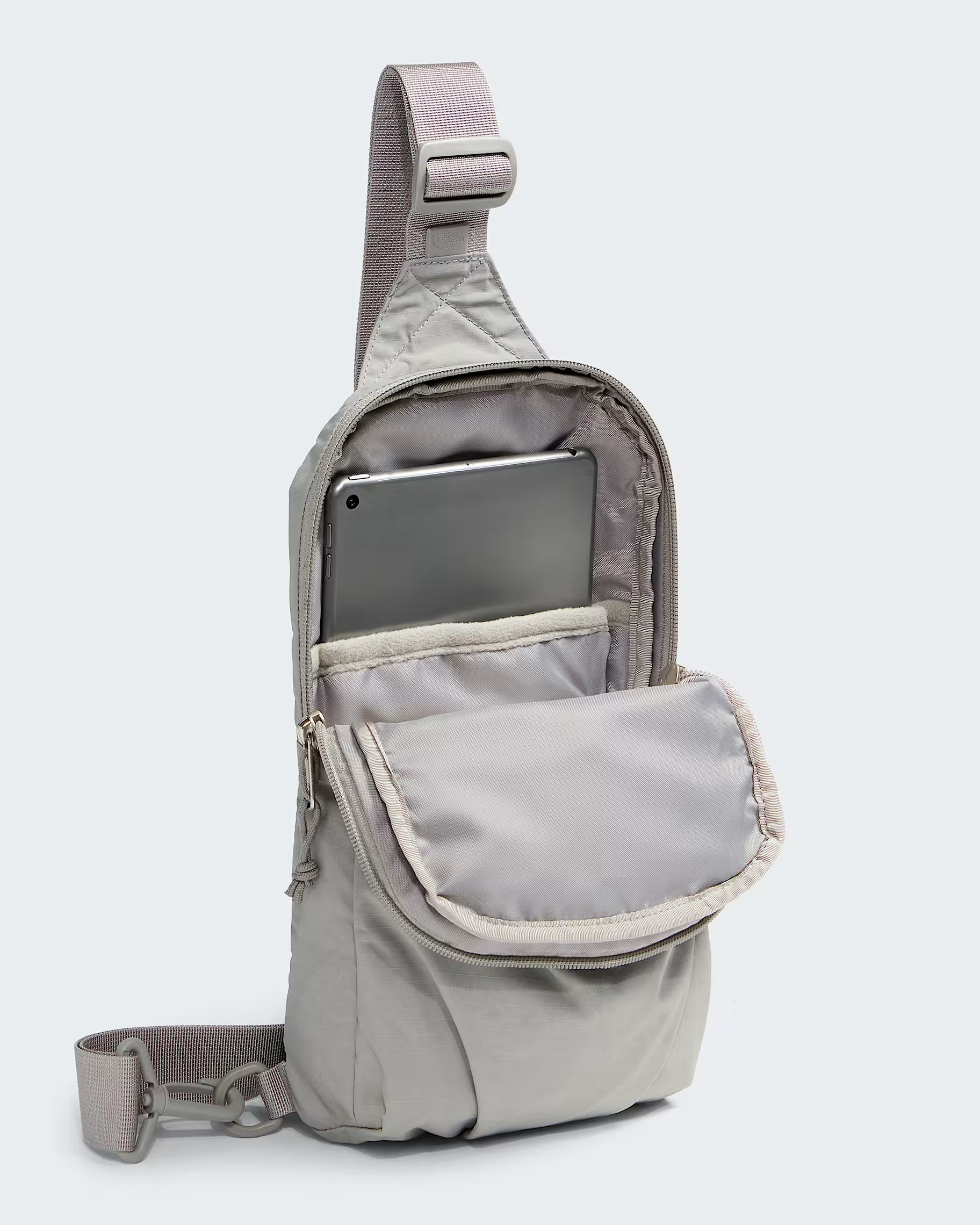 A light gray nylon sling backpack shown open, with a tablet inserted into the top compartment and a front flap pocket. The bag has a wide adjustable shoulder strap and a detachable crossbody strap, photographed on a clean light gray background.