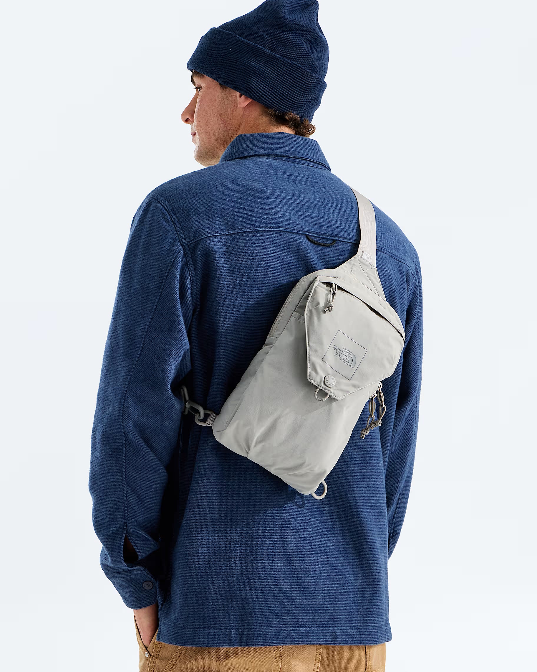 A man wearing a navy blue beanie, blue denim shirt, and tan pants is shown from the back, wearing a light gray sling backpack across his back. The photo is taken against a clean, light gray studio background.