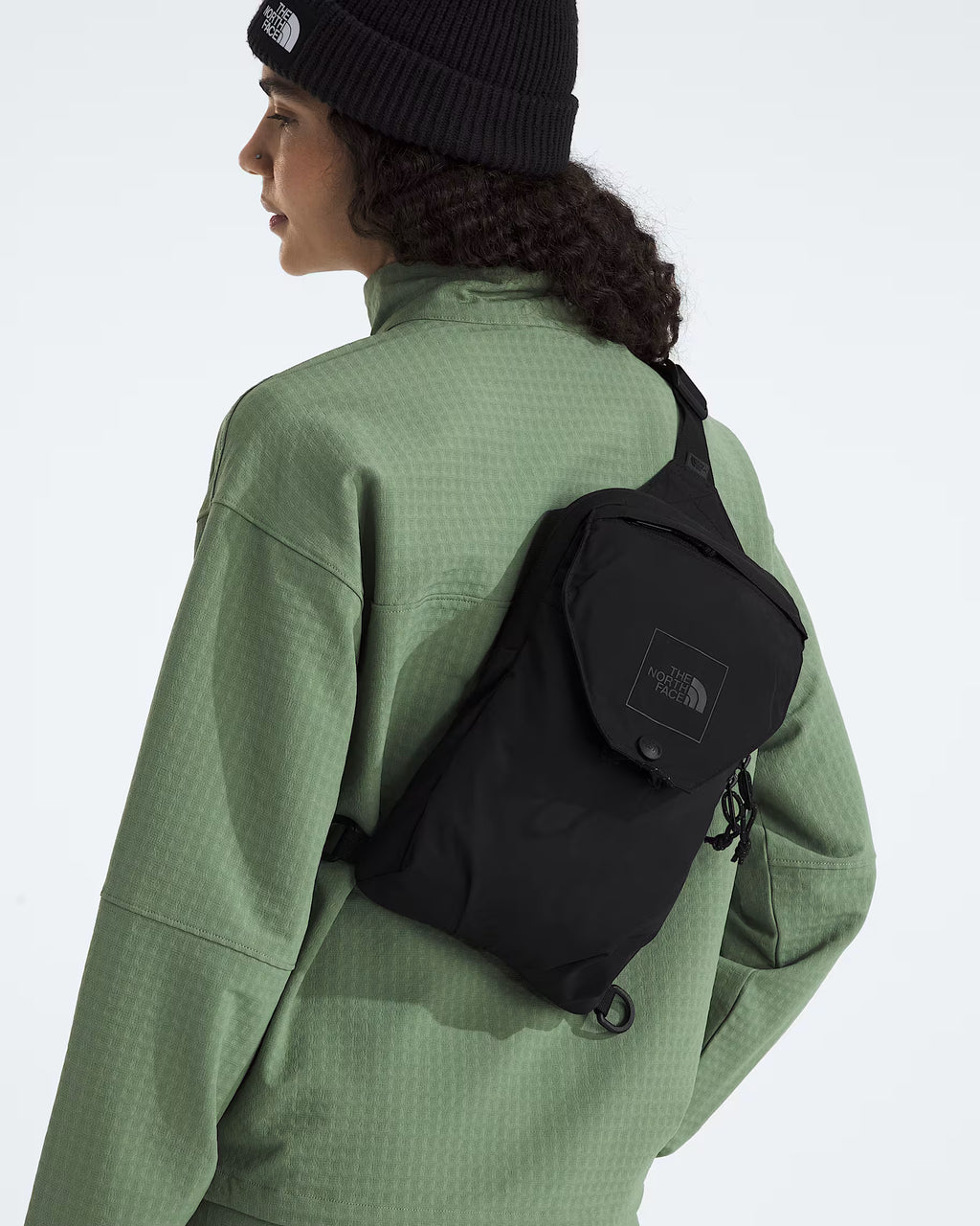 A person shown from the back wearing a black The North Face sling bag across their shoulder, paired with a sage green textured jacket and a black beanie.