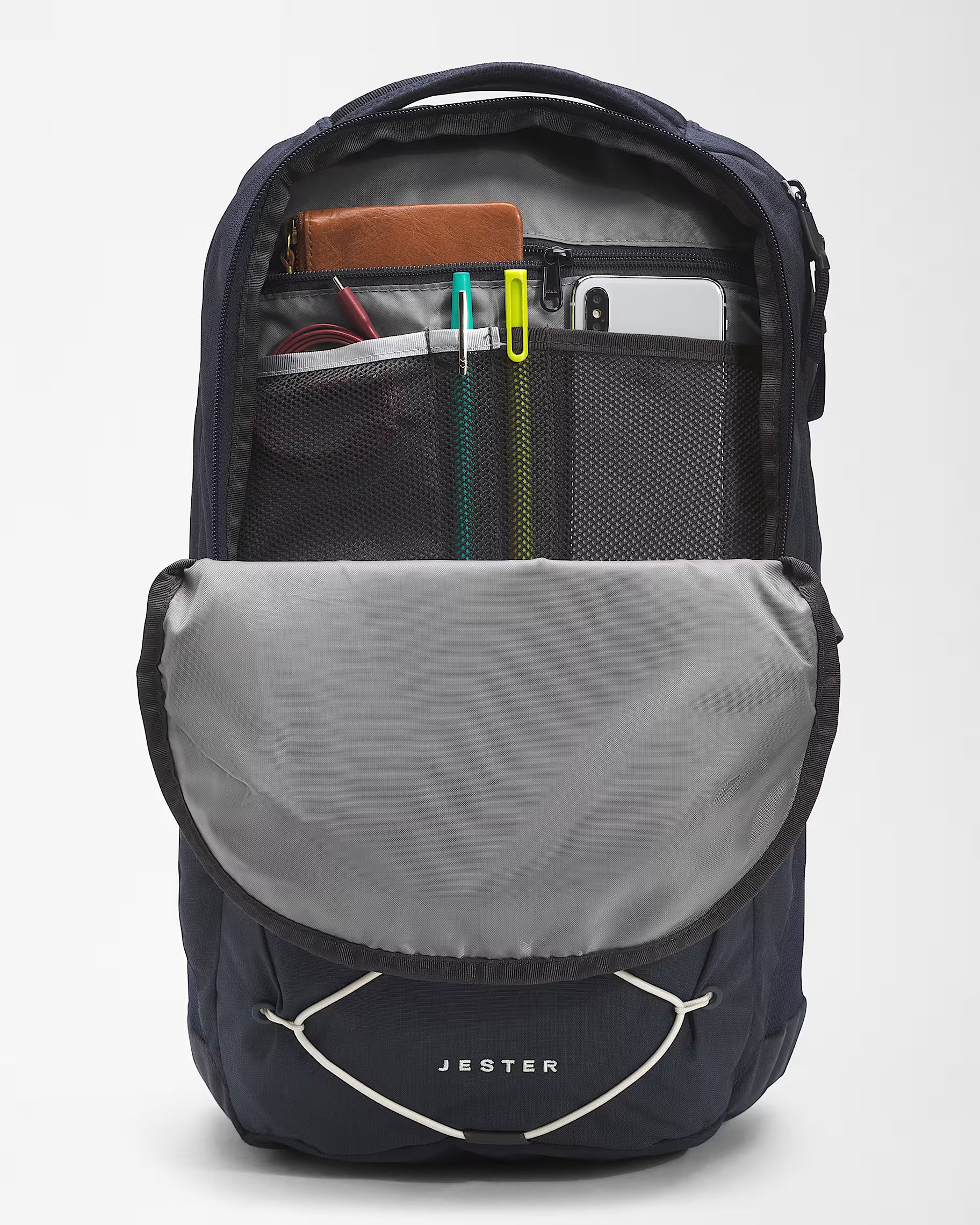 An open Jester backpack shown from the front, revealing a gray interior with mesh pockets holding a phone, tablet, and accessories. The exterior is dark navy blue with a light gray front flap and white Jester branding.