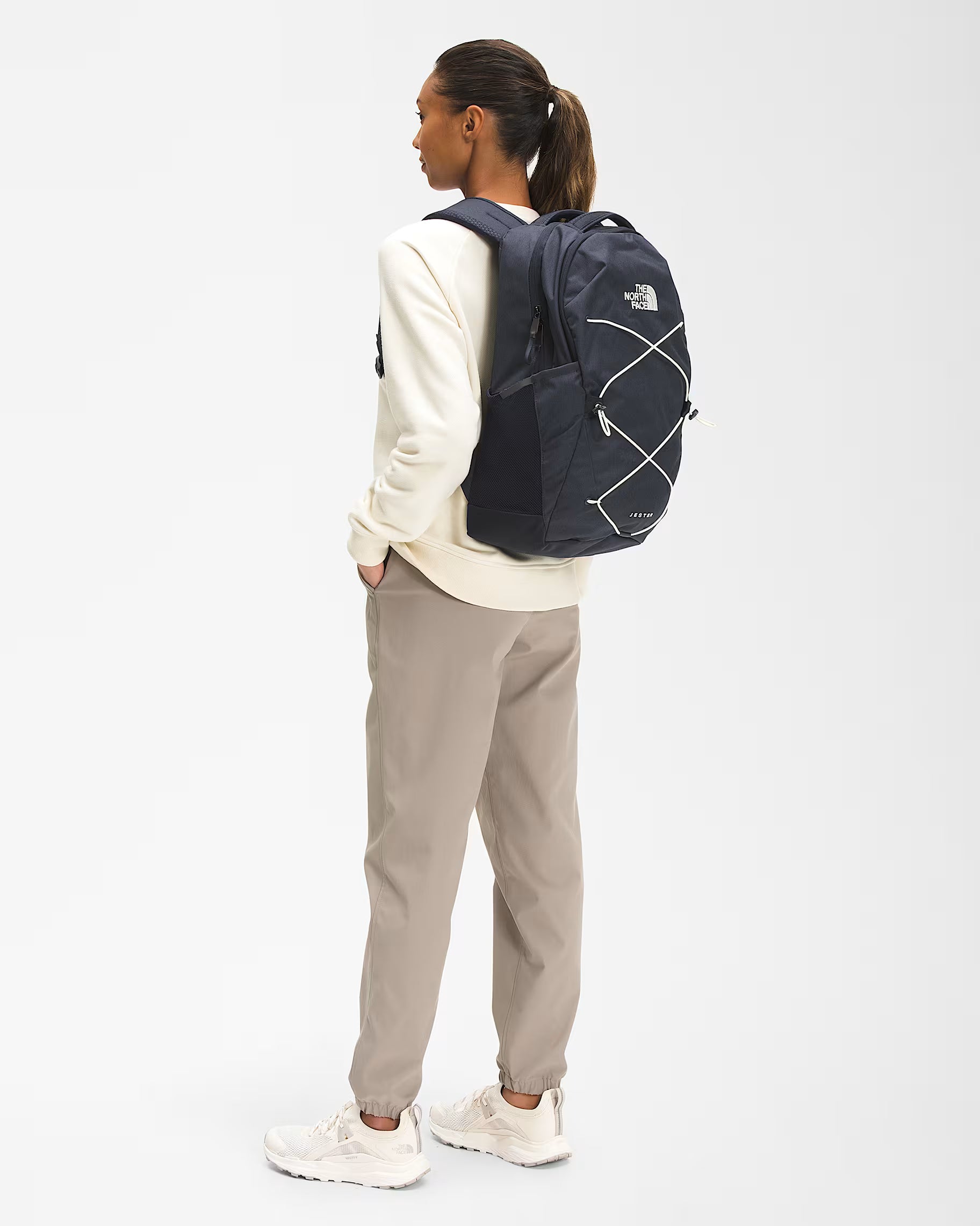 A woman shown from the back wearing a navy blue The North Face backpack, a cream sweatshirt, beige pants, and white sneakers against a plain white studio background.