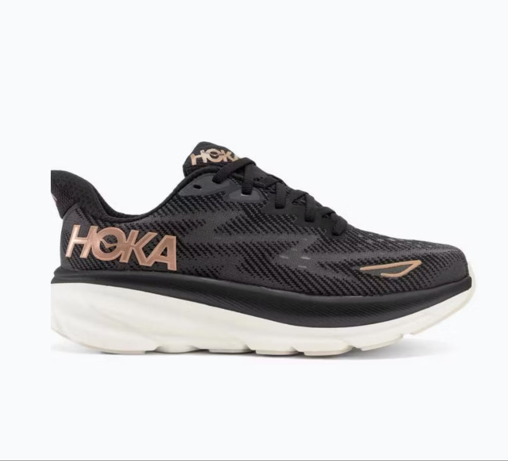 A Hoka running shoe in black with metallic rose gold accents on the logo and toe cap, set against a plain white background.