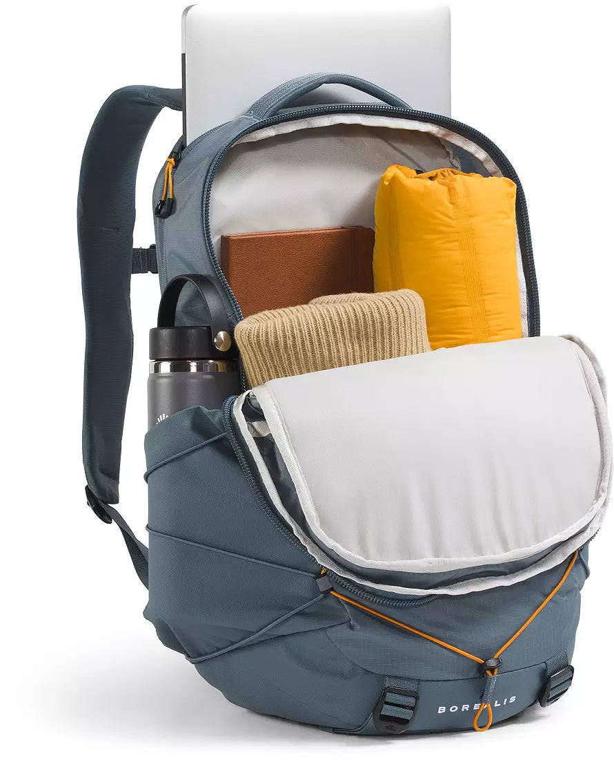 A slate blue-gray Borealis backpack shown open, filled with travel items including a yellow sleeping bag, a beige knit sweater, a brown book, a silver water bottle, and a white laptop. The backpack features a front flap pocket and side pocket, with orange zipper pulls and a visible Borealis logo on the bottom front.
