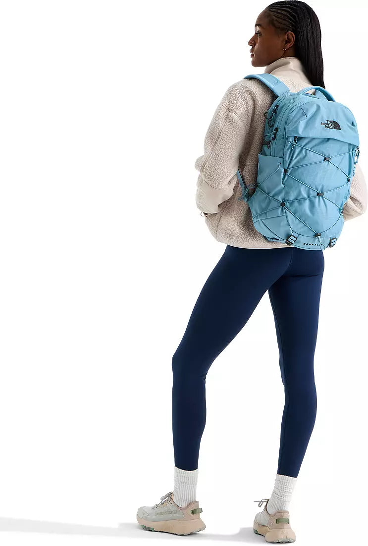 A woman wearing a cream knit sweater, navy leggings, white socks, and beige sneakers is shown from the back wearing a light blue quilted backpack by The North Face against a white studio background.