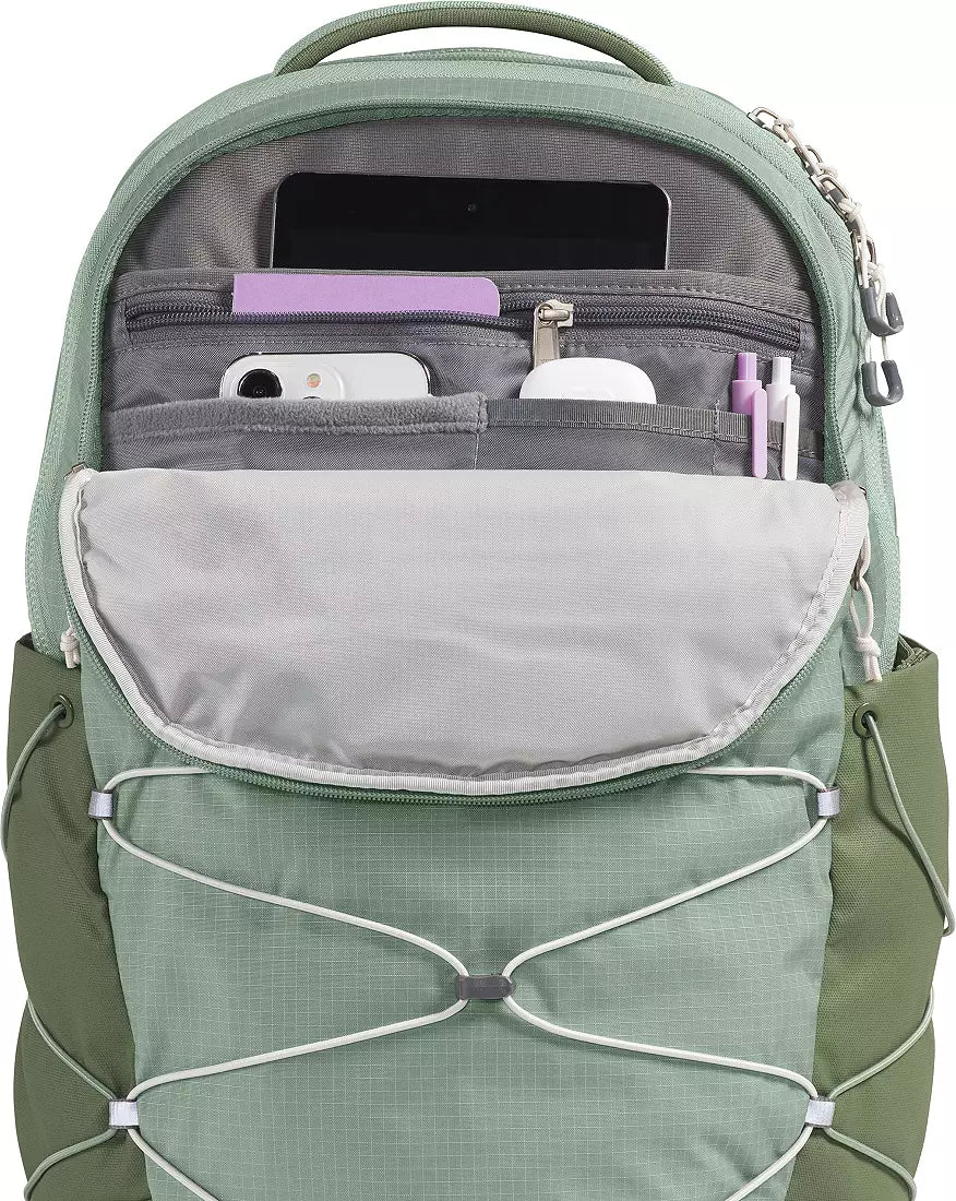 A sage green backpack shown open, displaying its organized interior compartments with a phone, wallet, and pens, on a clean white background.