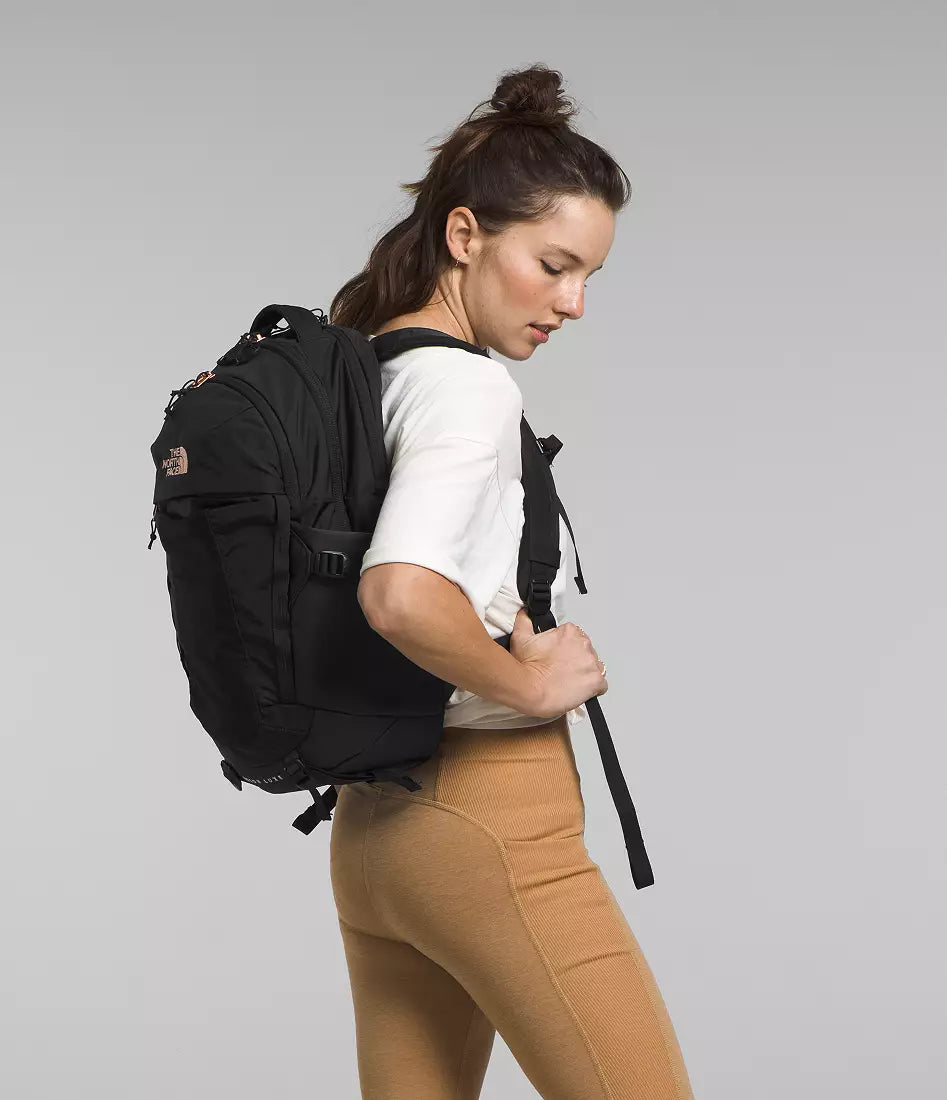 A woman wearing a black The North Face backpack, white t-shirt, and tan leggings, shown from a side-back angle against a solid gray studio background.