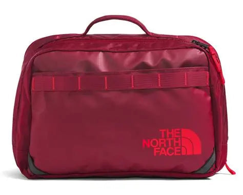A The North Face utility bag in a deep burgundy red, featuring a top carry handle, a horizontal webbing strap across the front, and the brand's logo printed in red on the lower right front.