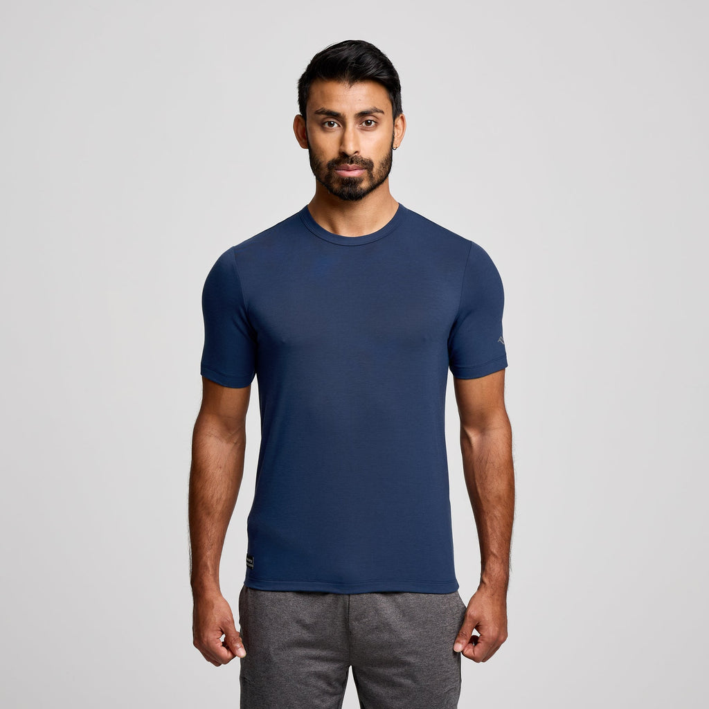 A man wearing a navy blue short-sleeve athletic t-shirt and gray sweatpants, standing against a light gray studio background.