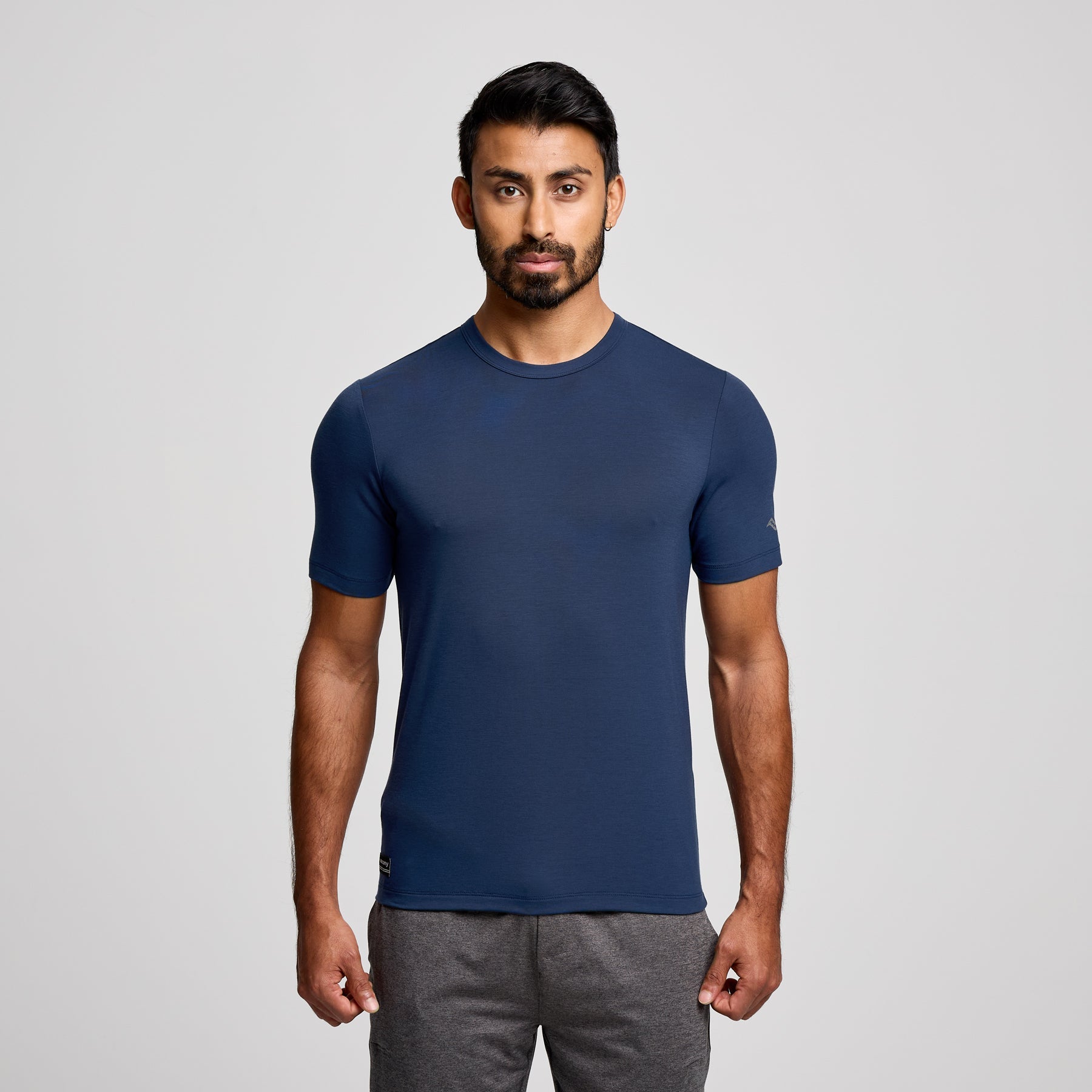 A man wearing a navy blue short-sleeve athletic t-shirt and gray sweatpants, standing against a light gray studio background.