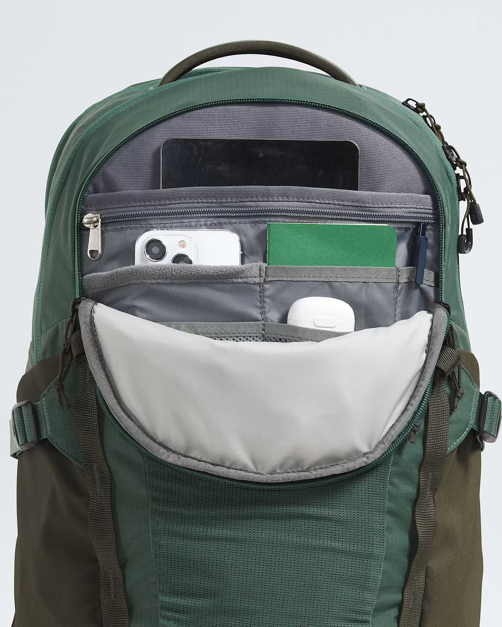 A close-up of a green backpack with its front pocket open, showing interior compartments holding a tablet, phone, card holder, and small accessories against a light gray background.
