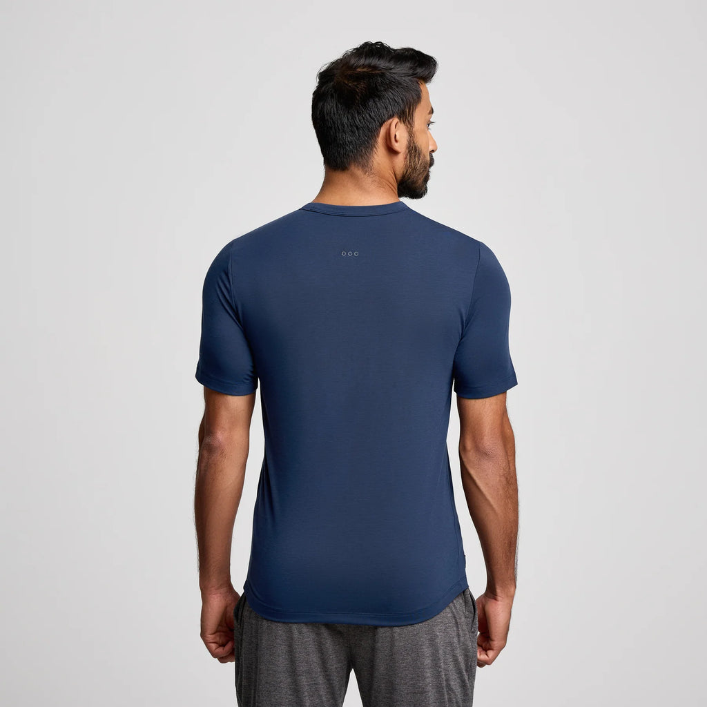 A man viewed from the back wearing a navy blue athletic t-shirt and gray athletic shorts against a light gray studio background.