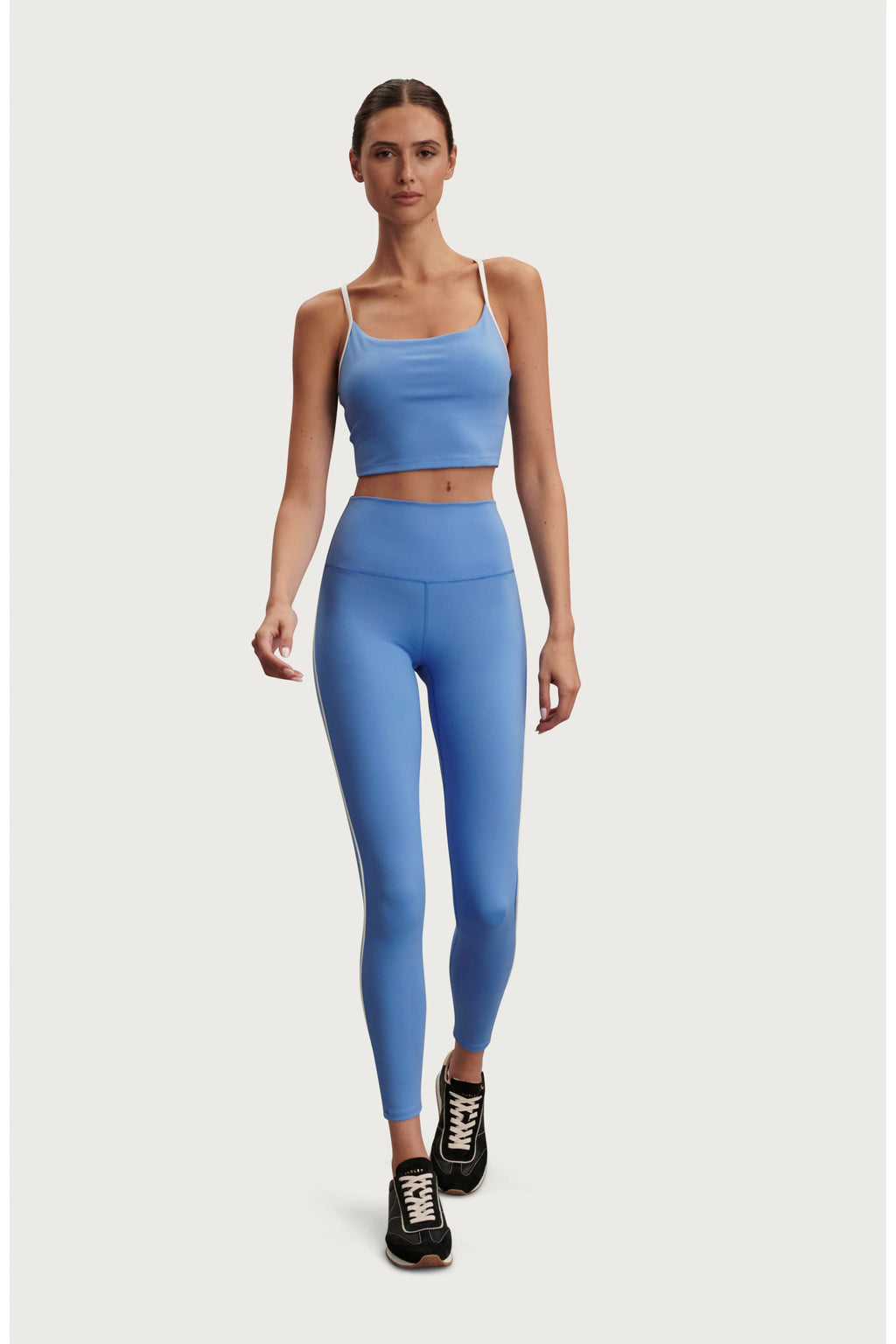 A woman modeling a matching light blue activewear set consisting of a spaghetti-strap crop top and high-waisted leggings, paired with black athletic sneakers, against a solid light gray background.