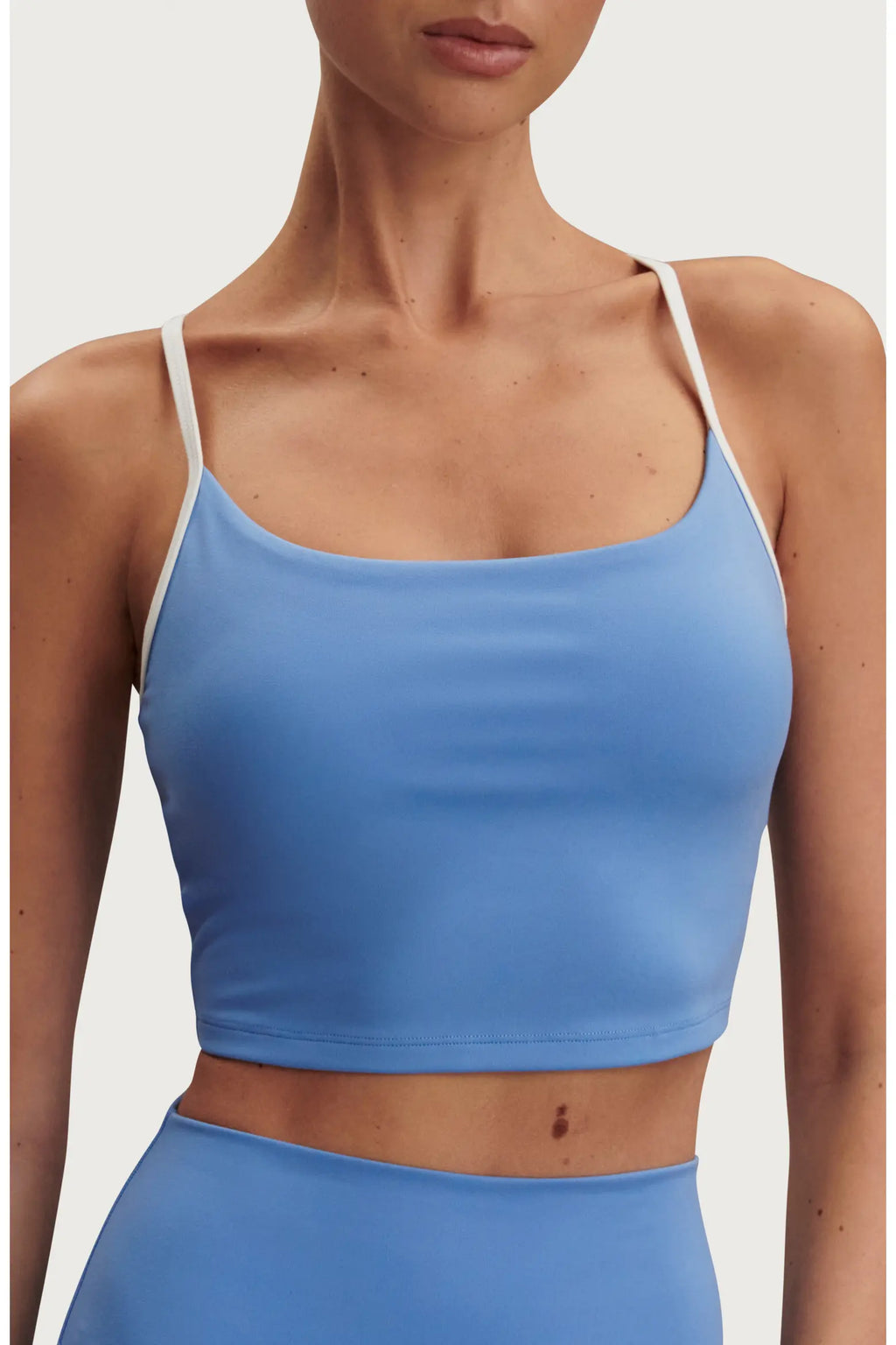A close-up of a model wearing a light blue crop top with thin white shoulder straps, paired with matching light blue bottoms, against a plain light background.