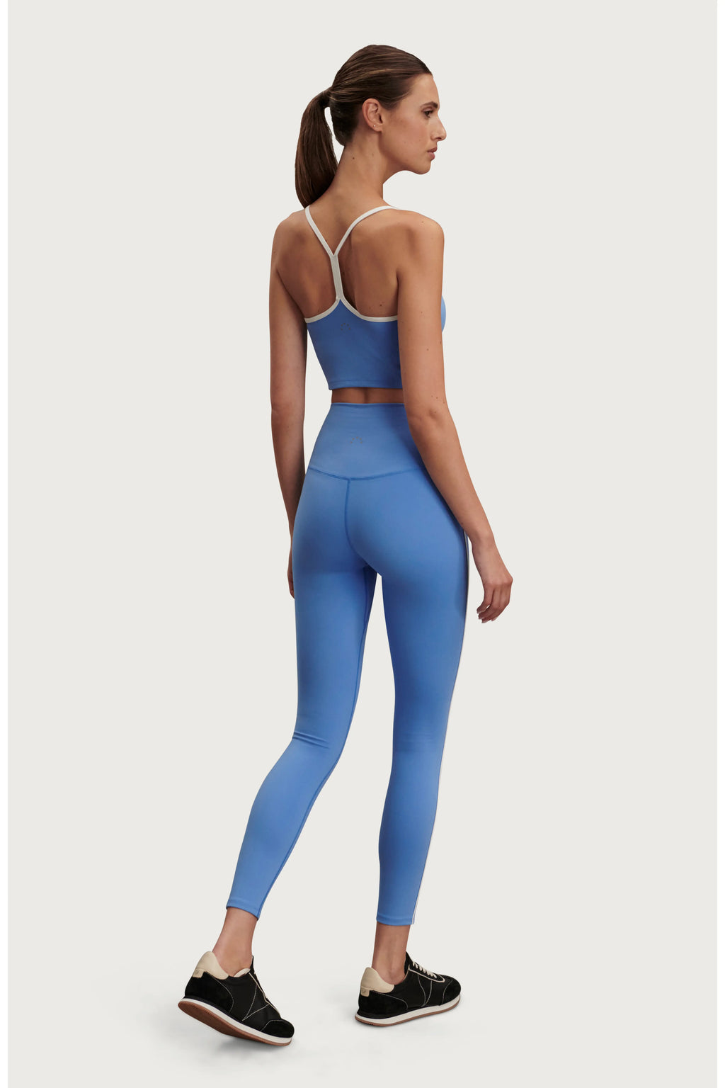 A woman shown from the back wearing a matching athletic set consisting of a blue sports bra and high-waisted leggings, paired with black sneakers, against a light gray studio background.