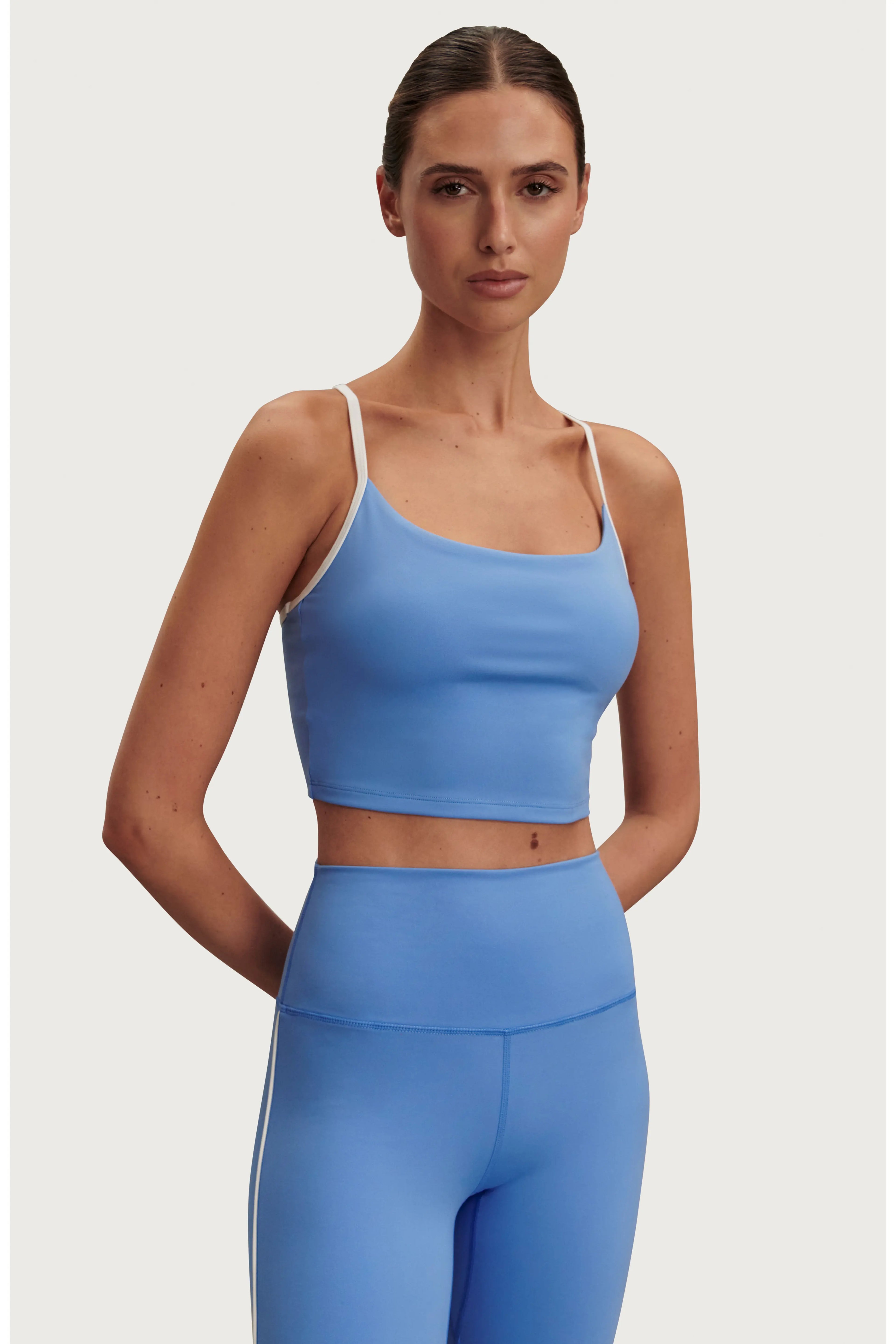 A woman modeling a light blue activewear set consisting of a scoop-neck crop top with thin white straps and matching high-waisted leggings, set against a solid light gray background.