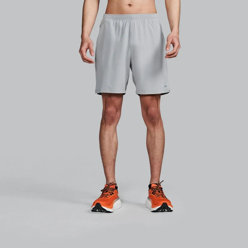 A model from the waist down wearing light gray athletic shorts and orange running shoes, standing against a solid gray studio background.